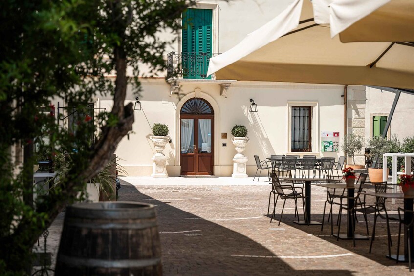 Un café al aire libre con mesas y sillas, un edificio con un persiano verde, y un árbol en primer plano. 4