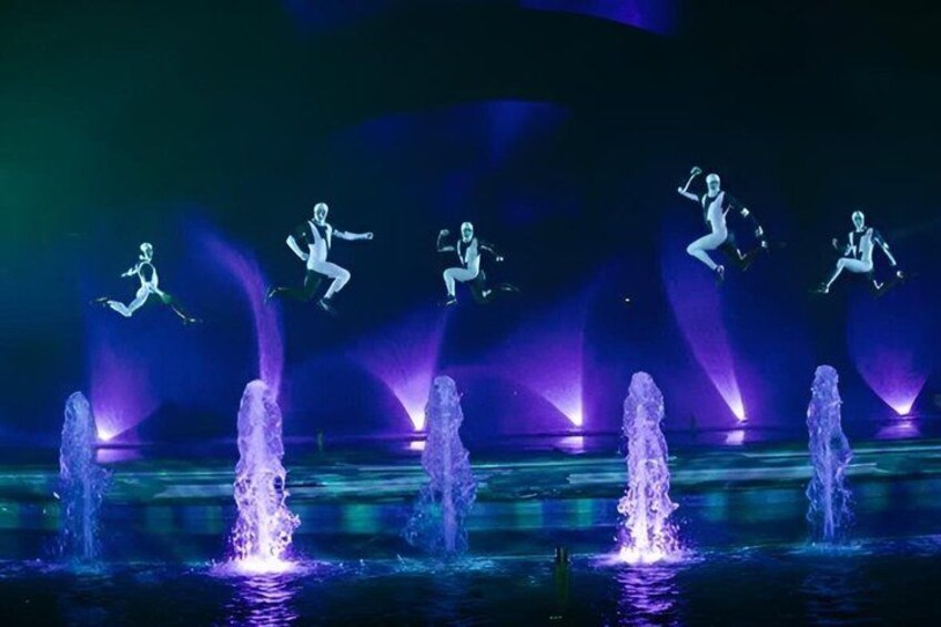 A water fountain with multiple jets, illuminated by purple lights, and a person jumping in the air. 4