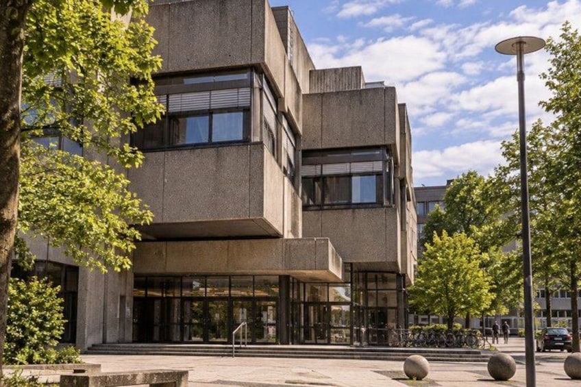 A modern building with a concrete facade, large windows, and a paved area with trees and bollards. 5