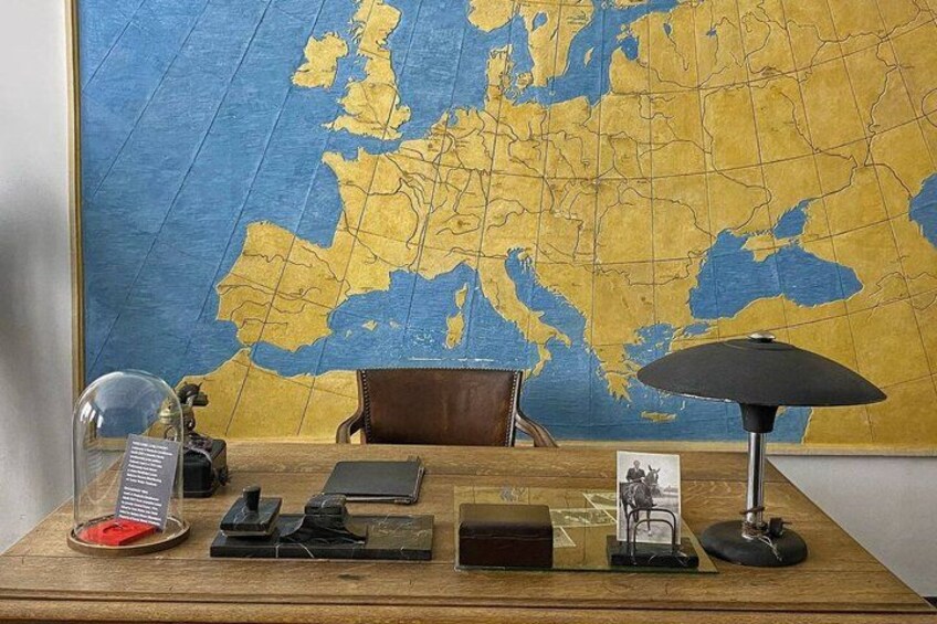A wooden desk with a large world map, a lamp, a leather chair, and various office items. 1