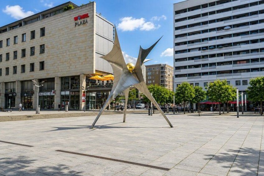 A modern sculpture in an open plaza with buildings in the background. 2