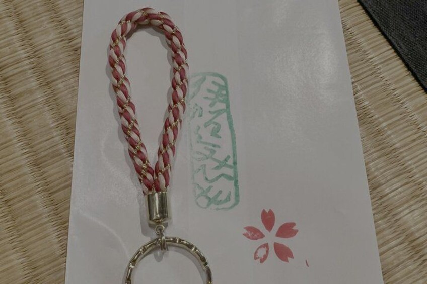 A keychain with a braided handle and a metal ring, a piece of paper with a red flower and some text, and a textured surface beneath. 3