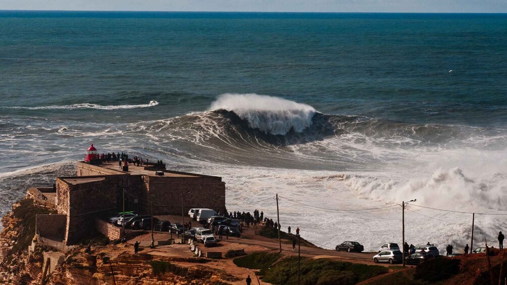 Escena costera con una ola grande rompiendo en la orilla, un faro y un grupo de personas observando el océano. 1
