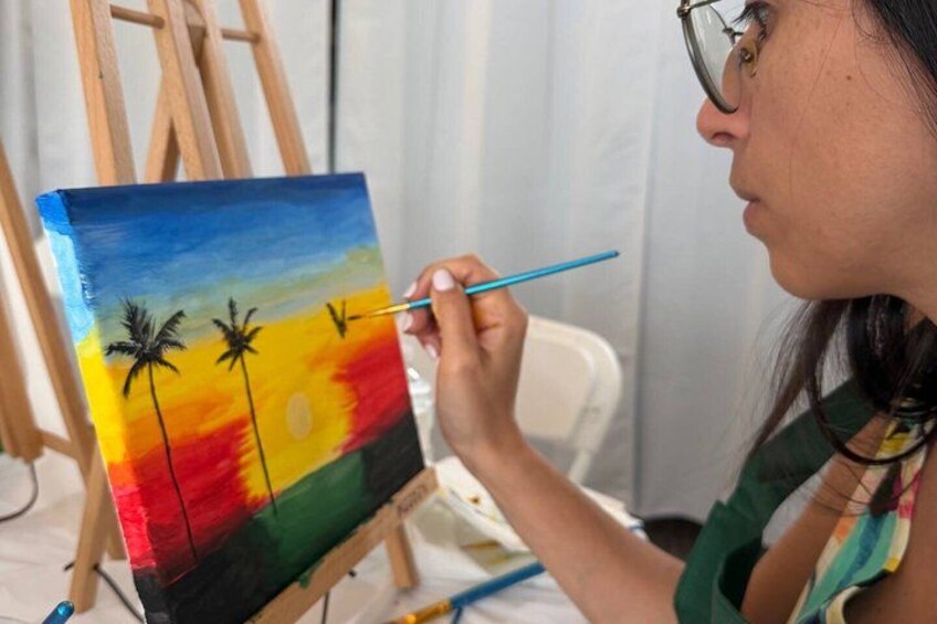 A person painting a sunset scene with palm trees on an easel. 4