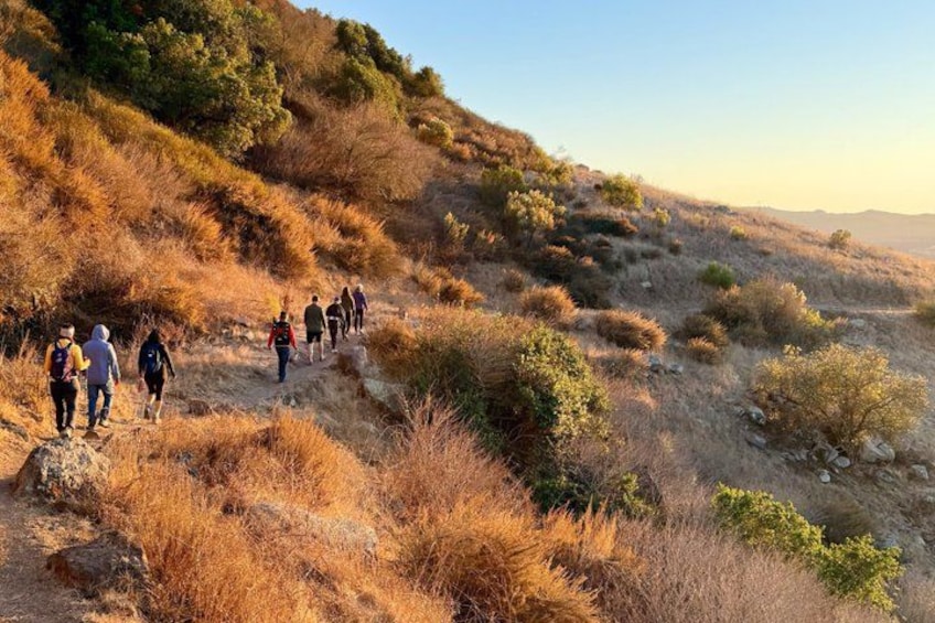 A group of hikers walking on a dry, grassy trail with sparse vegetation and hills in the background. 4