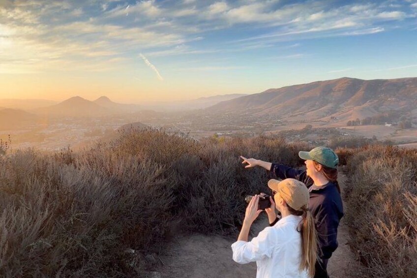 Two people hiking on a trail with a view of a valley and mountains in the background. 1