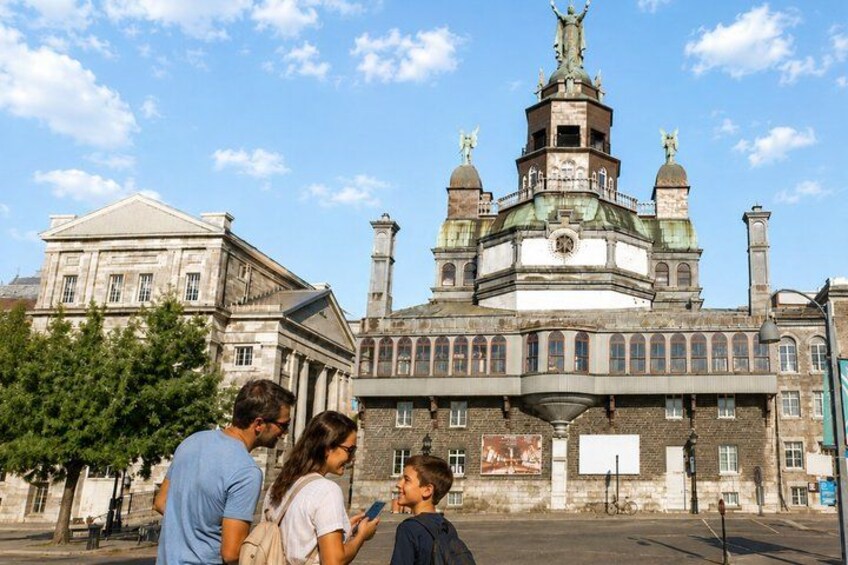 A historic building with a central tower, a large open plaza, and a few people standing in the foreground. 1