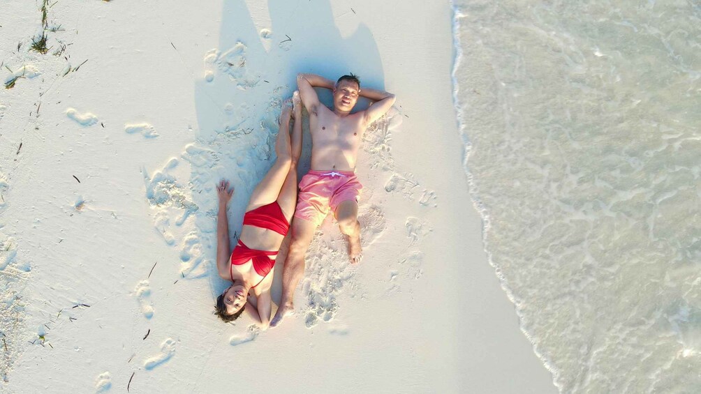 Deux personnes allongées sur une plage de sable, avec des empreintes de pas autour d’elles. 2