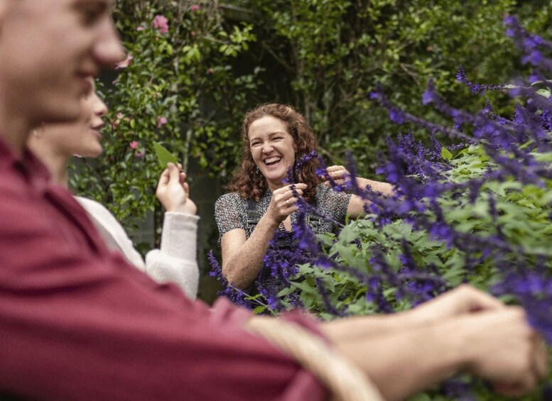 A person holding purple flowers in a garden with greenery and pink flowers in the background. 2