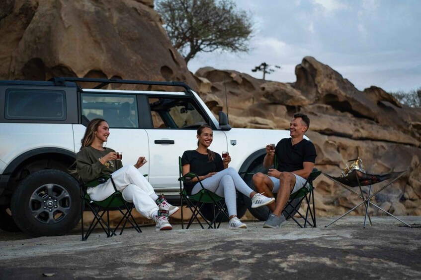 Trois personnes sont assises sur des chaises devant un véhicule blanc, avec un paysage rocheux en arrière-plan. 2