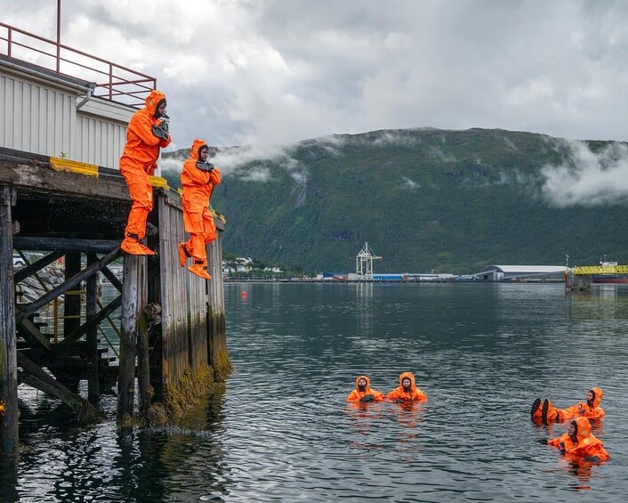 Des personnes vêtues de combinaisons orange se trouvent sur un quai, certaines debout et d’autres dans l’eau. On aperçoit une montagne et des structures industrielles en arrière-plan. 3