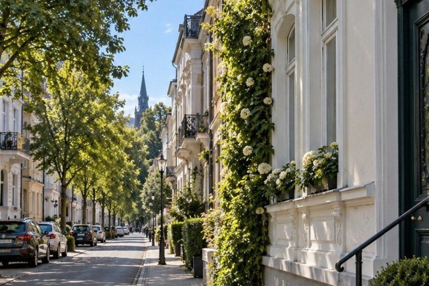 A tree-lined street with parked cars and buildings with white facades and balconies. 4
