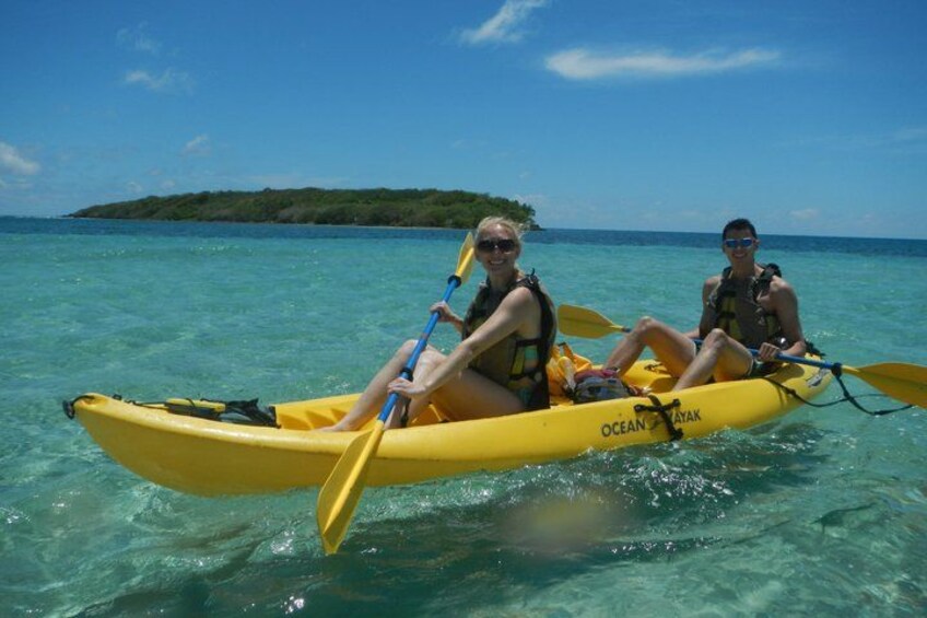 Two people kayaking on clear turquoise water with a small island in the background. 1