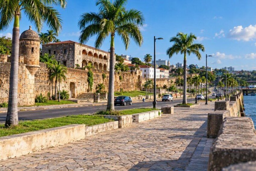 A coastal cityscape with historic stone buildings, palm trees, and a paved walkway along the waterfront. 3
