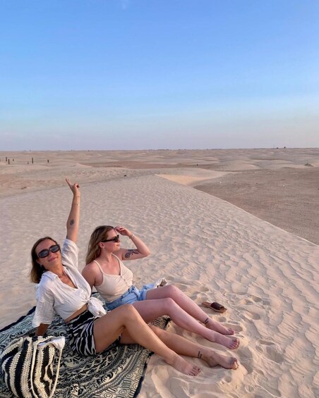Deux personnes sont assises sur une couverture à motifs, dans un paysage désertique parsemé de dunes de sable. 5