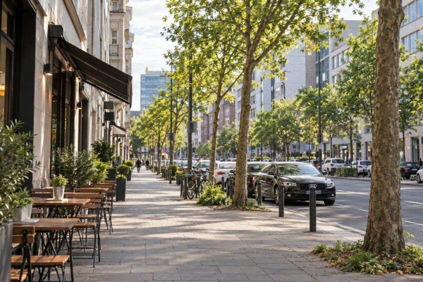 A tree-lined sidewalk with outdoor seating, parked cars, and buildings. 3