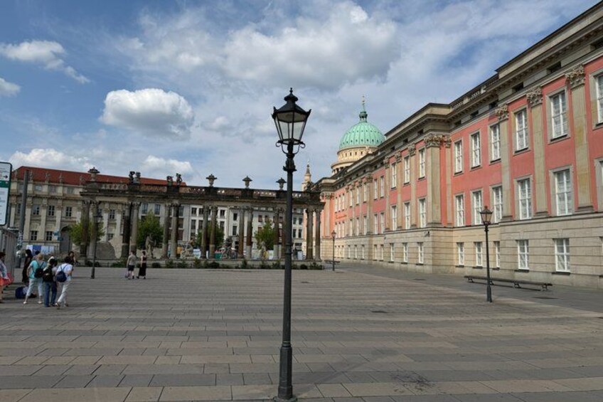 A spacious square with a historic building featuring a dome and columns, a street lamp, and a few people walking. 3