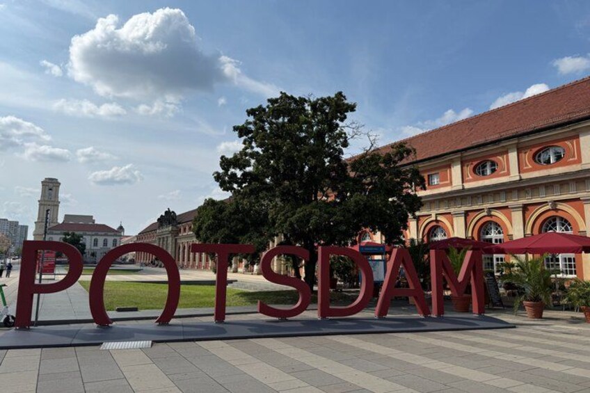 A large red sign with the word 'POTSDAM' in the foreground, a building with arched windows in the background, and a clear sky with clouds. 4