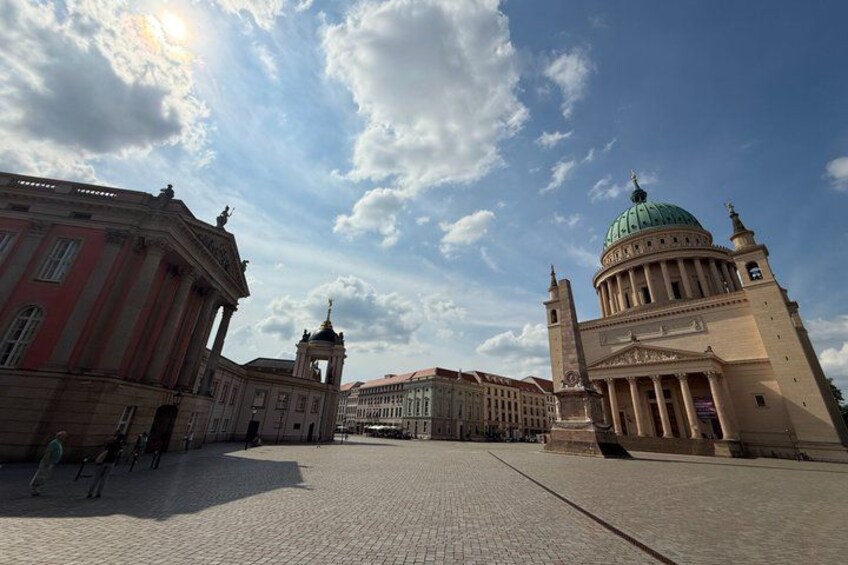 A historical square with classical architecture, including a prominent building with a green dome and a red building with columns. 2