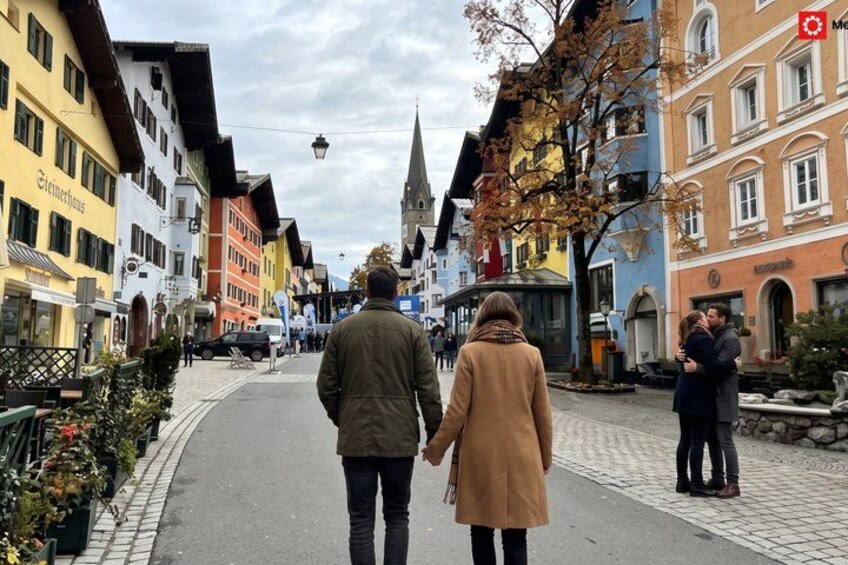 A couple walking down a cobblestone street lined with colorful buildings, a church spire in the distance, and a small fountain. 2