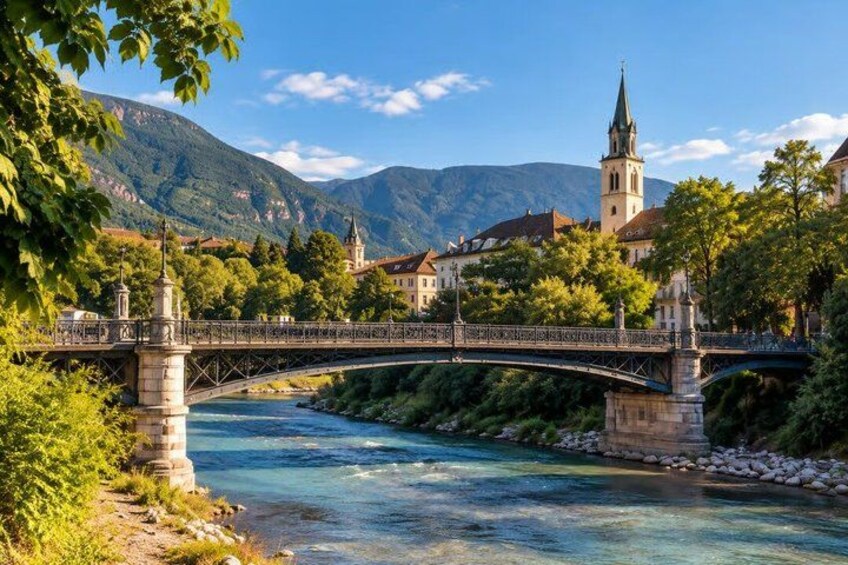 A stone bridge over a river, a church with a tall spire, and mountains in the background. 4