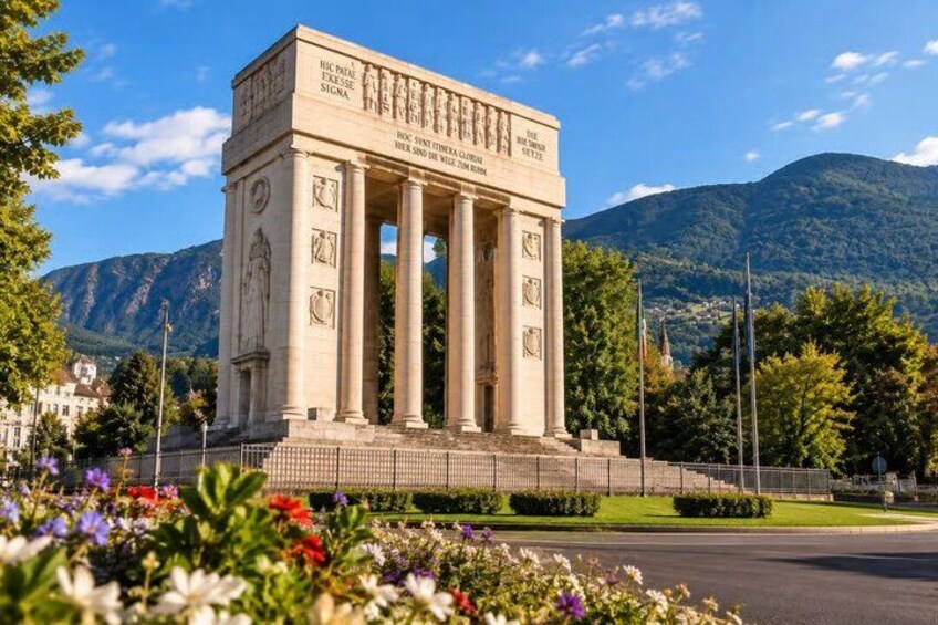 A neoclassical monument with columns, surrounded by a well-maintained garden with various flowers, set against a backdrop of mountains and a clear sky. 5