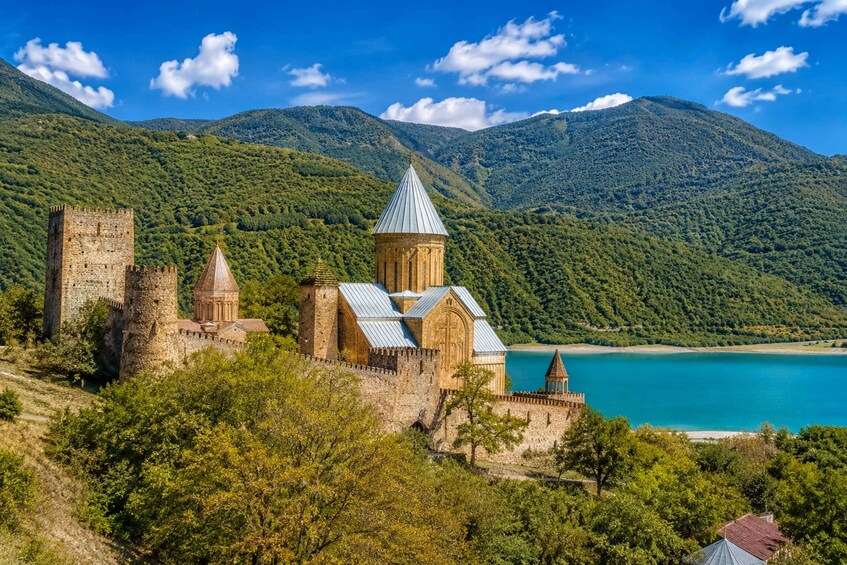 A historic castle with multiple towers and a church with a pointed roof, surrounded by lush green hills and a clear blue lake. 4