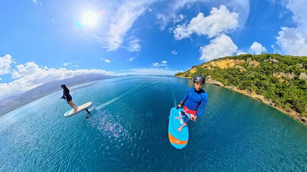 Two people surfing on a clear blue sea with a mountainous coastline in the background. 1