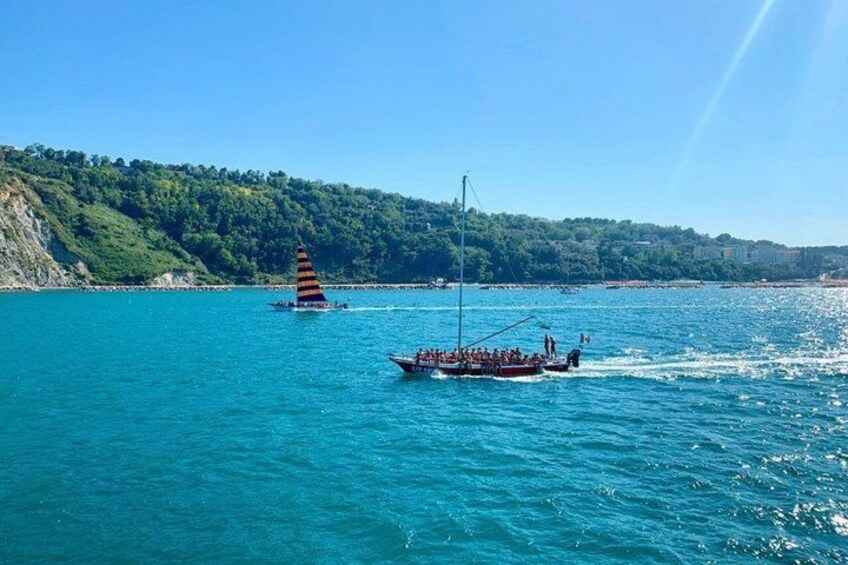 A boat with people on a calm sea, a sailboat with a striped sail, and a forested coastline. 2