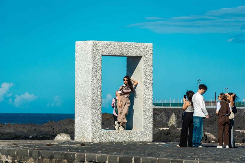 A group of people standing near a large, textured concrete structure with a blue-tinted window, overlooking a coastal landscape with the sea in the background. 1