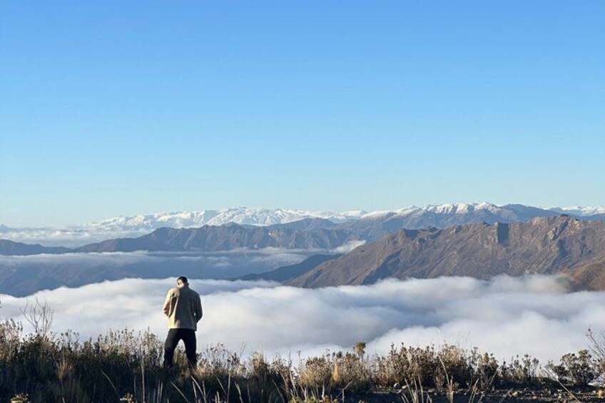 A person standing on a grassy hill overlooking a mountain range with snow-capped peaks and a layer of clouds below. 2