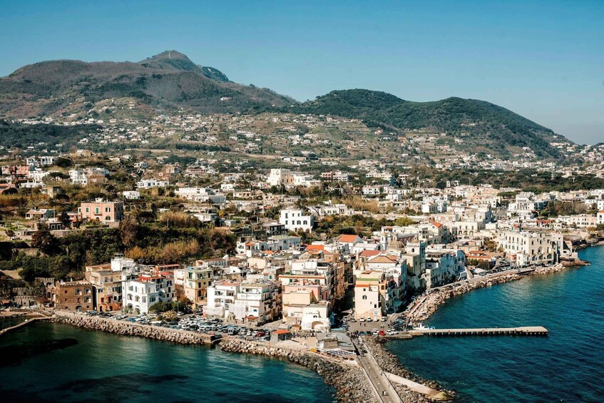 A coastal town with buildings along the waterfront, a rocky breakwater, and hills in the background. 2