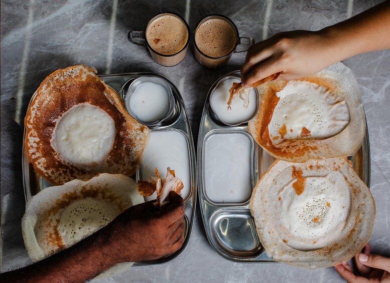 Una bandeja con dos platos de dosa, dos tazas de café y una mano alcanzando un trozo de dosa. 4