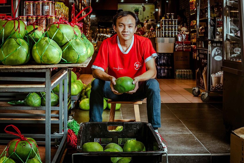 Una persona sentada en un taburete rodeada de cocos verdes, algunos atados con cintas rojas, en un mercado. 2