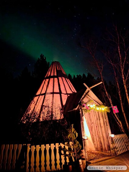 A wooden structure with a conical roof, a small shed, and a fence under a night sky with the aurora borealis. 3