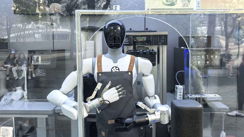 Un maniquí de robot humanoide con delantal negro y casco, ubicado frente a una máquina expendedora y una vitrina de vidrio. 2