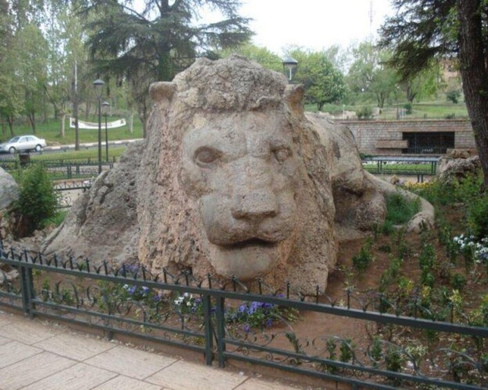 Une grande sculpture en pierre se trouve dans un parc, avec une clôture métallique en premier plan. 3