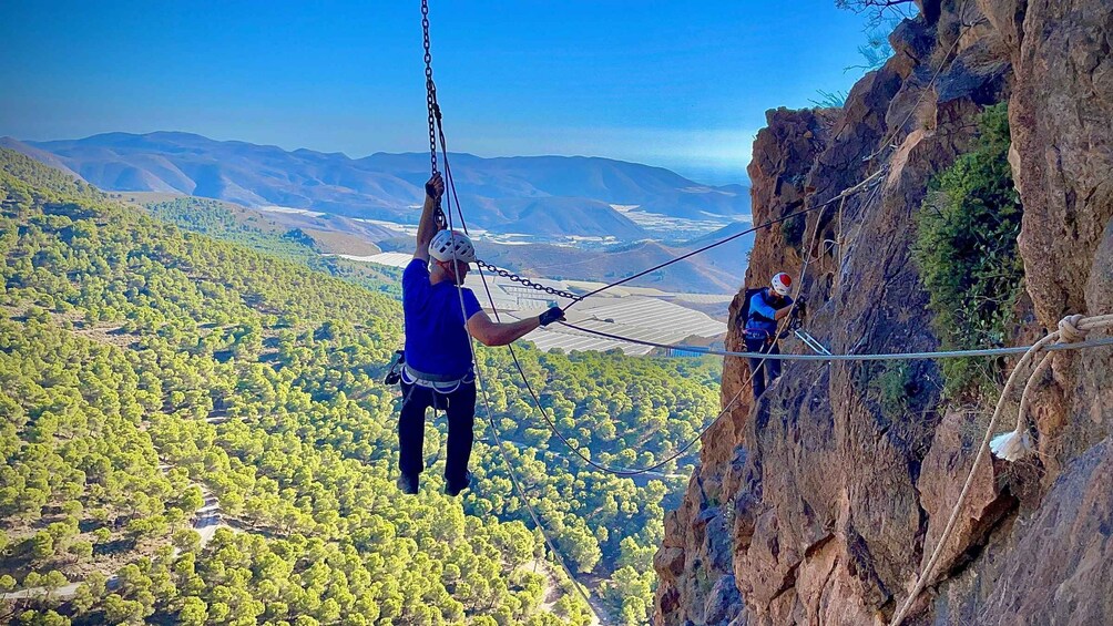 A person rappelling down a cliff with a rope, another person assisting, and a vast landscape with trees and distant mountains. 4