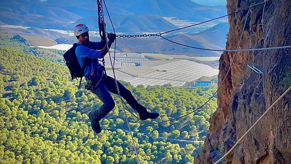 A person rappelling down a cliff with a harness and helmet, surrounded by a forested landscape and distant buildings. 1