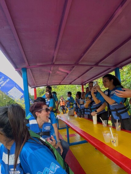 Un grupo de personas con chalecos salvavidas azules sentadas en una mesa bajo una zona techada con techo rojo. 5