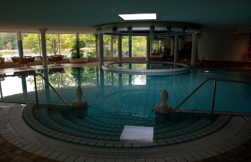 An indoor swimming pool with a tiled floor, a central fountain, and surrounding columns. 2