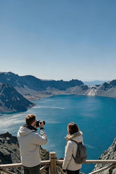 Two people taking photographs of a lake surrounded by mountains. 1
