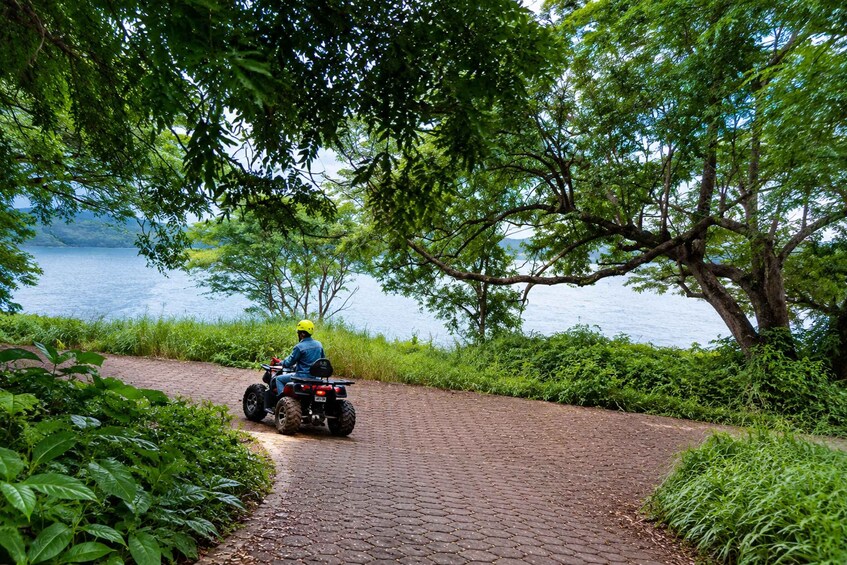 A person riding an ATV on a paved path surrounded by lush greenery and a body of water. 2