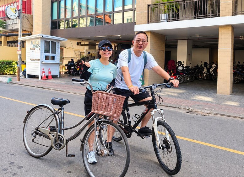 Dos personas en una bicicleta tándem con canasta, estacionada en una calle con edificios y motocicletas aparcadas al fondo. 3
