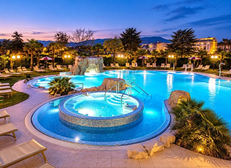 A swimming pool with a hot tub, lounge chairs, and a waterfall feature at dusk. 3