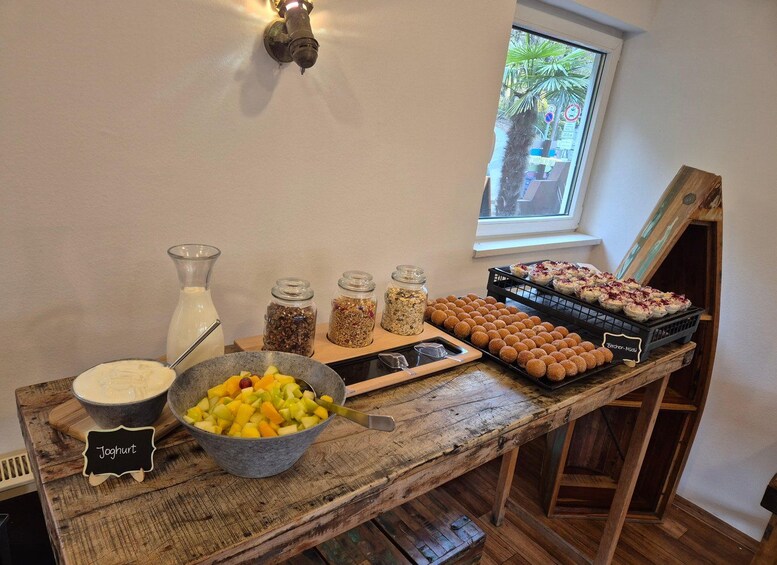 A wooden table with food items, including a bowl of fruit, jars of granola, and trays of pastries. 5