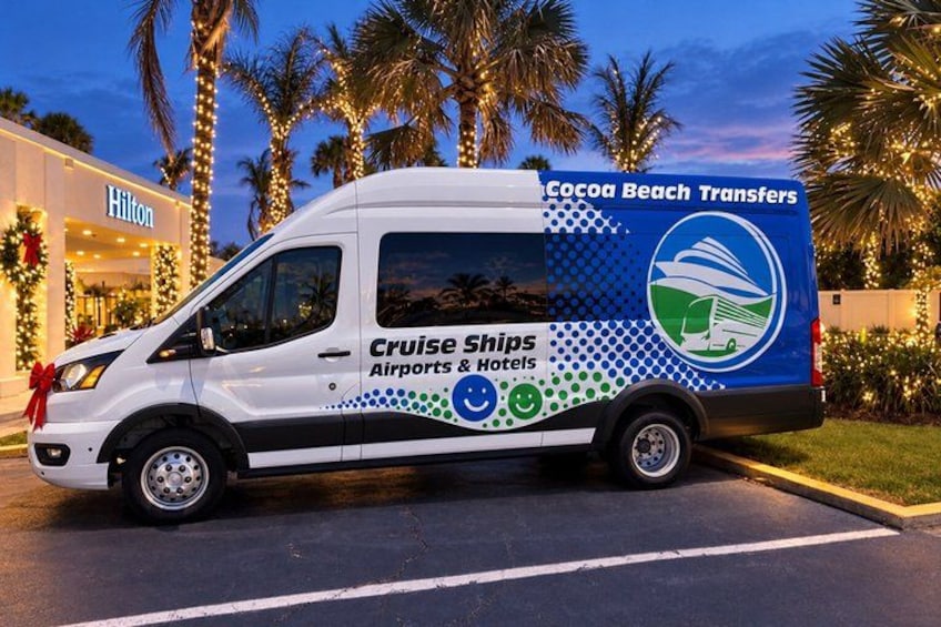 A white van with 'Cruise Ships Airports & Hotels' and 'Cocoa Beach Transfers' branding parked in front of a Hilton hotel. 2