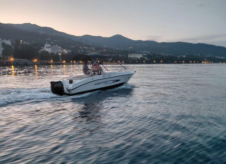 A white motorboat with two people on it, moving on calm water with a cityscape and mountains in the background. 2