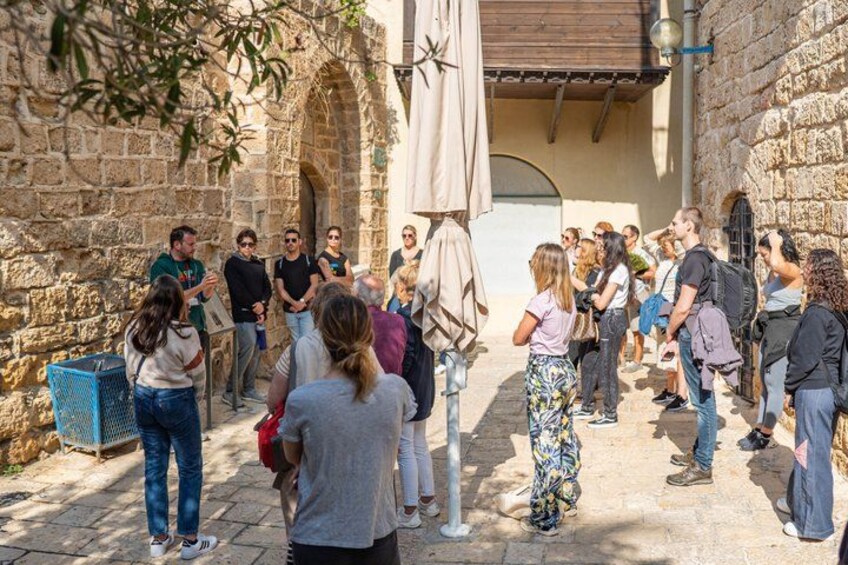 A group of people gathered in a stone courtyard with arches, a blue trash bin, and a white umbrella. 5
