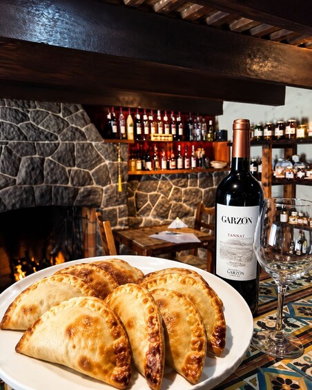 A plate of empanadas, a bottle of Garzon Tannat wine, and a glass of wine on a table with a stone fireplace in the background. 1